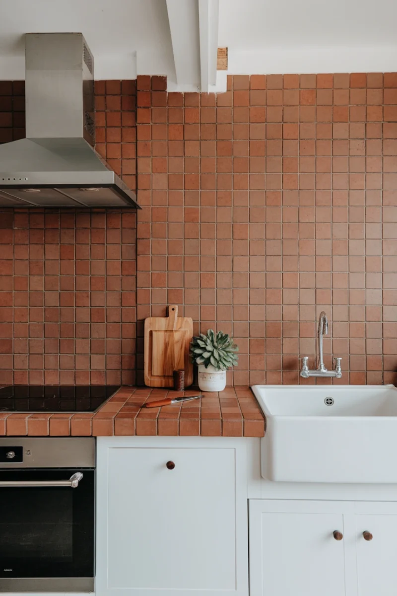 A modern kitchen featuring earthy terracotta wall tiles, a white sink, and wooden accents.